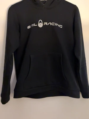 Svart hoodie från Sail Racing - Svart hoodie från Sail Racing med vit logga tryckt över bröstet. Klassisk känguruficka framtill och huva. Tillverkad i mjukt material som passar perfekt för en avslappnad stil. Snygg och enkel design som funkar till det mesta.