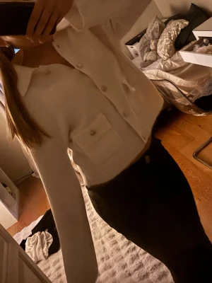 Vit kofta med fickor och knappar - Säljer en stilren vit kofta med stora knappar framtill och två praktiska fickor på magen. Koftan har en klassisk krage och är gjord i ett mjukt, stickat material. Perfekt att slänga på sig för en clean och enkel look.