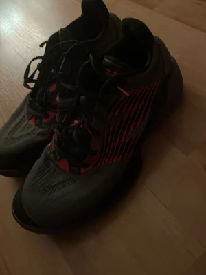 New balance basketskor - Säljer ett par gråa New Balance sneakers med svarta och röda detaljer. Skorna har lågt skaft, snörning och en mönstrad sula för bra grepp. Perfekta för dig som gillar sportig stil och vill ha något bekvämt till vardags.