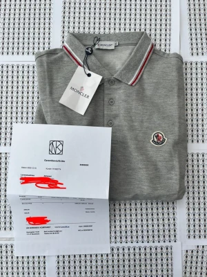 Grå Moncler pikétröja med röd/vit kant - Snygg grå pikétröja från Moncler med klassisk krage och knappar framtill. Tröjan har röda och vita detaljer på krage och ärmslut samt Moncler-logga på bröstet. Perfekt för en clean och sportig look.