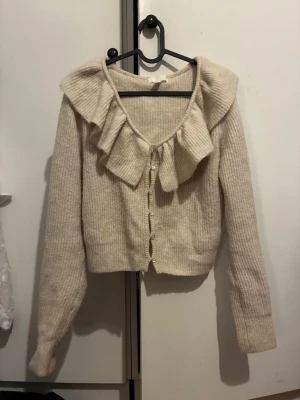 Beige kofta med volangkrage - Supersöt beige kofta med stora volanger runt ringningen och knappar framtill.