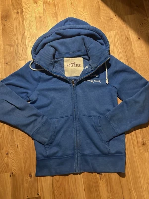 Blå sällsynt zip-hoodie från Hollister - Snygg sällsynt blå hoodie från Hollister med dragkedja och vit broderad logga på bröstet. Mjuk insida i fleece, två fickor framtill och justerbar huva med vita snören.