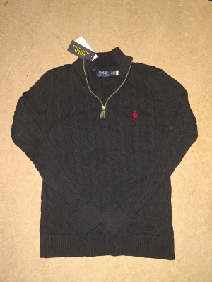 Svart kabelstickad tröja Polo Ralph Lauren - Kabelstickad svart tröja från Polo Ralph Lauren med halv dragkedja och ståkrage. Tröjan har det klassiska röda Polo-logot broderat på bröstet och ribbade muddar vid ärmslut och nederkant. Perfekt för lager på lager och snygg till jeans.