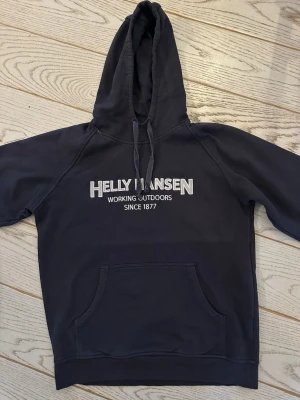 Mörkblå Helly Hansen hoodie - Snygg mörkblå hoodie från Helly Hansen med stor magficka och dragsko i huvan. Tryck på bröstet med texten 'Helly Hansen Working Outdoors Since 1877'. Perfekt för chill dagar eller när du vill ha en sportig look.