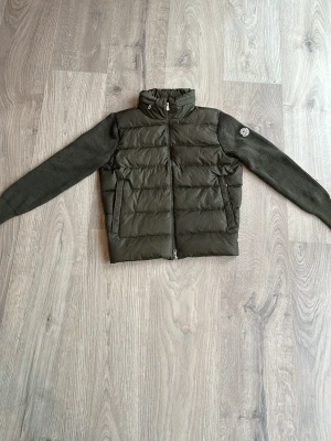 Grön dunväst från Moncler - Snygg grön dunväst från Moncler med stickade ärmar och quiltad kropp. Västen har hög krage, dragkedja framtill och Moncler-logga på ena ärmen. Perfekt för lager-på-lager och ger en sportig vibe. Helt ny. Alla taggar etiketter funkar och ingår. Storlek L men lutar även mot M hållet. 