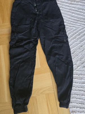 Svarta cargobyxor med resår - Säljer ett par svarta cargobyxor med flera stora fickor och resår i bensluten. Byxorna har en avslappnad passform och dragsko i midjan. Perfekta för en streetwear-look och riktigt sköna att bära till vardags. Något korta för mig som är 170cm lång 