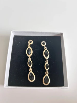 Guldiga örhängen med svarta stenar - Orginalpris 1000 och köpte för bröllop. Använt en gång. Snygga hängande örhängen i guldton med tre svarta, facetterade stenar i varje örhänge. Stenarna är infattade i droppformade och marquiseformade ramar. Perfekta för dig som gillar stilrena accessoarer med en touch av elegans.