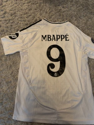 Real Madrid Mbappé matchtröja - Säljer en vit Real Madrid matchtröja från Adidas med Mbappé och nummer 9 på ryggen. Tröjan har svarta detaljer, klubbmärke på bröstet och sponsortryck 'Emirates Fly Better' framtill. Klassisk sportdesign med korta ärmar och Aeroready-material.