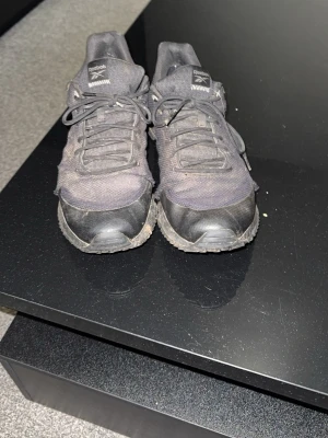 Svarta Reebok sneakers i textil - Säljer ett par svarta Reebok sneakers med lågt skaft. Skorna har ovandel i mesh och syntet, klassisk snörning och detaljer i gummi på tån och hälen. Perfekta för dig som gillar sportig stil och vill ha bekväma skor till vardags.