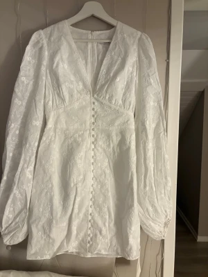Vit kort klänning med spetsdetaljer - Säljer en vit kort klänning med v-ringning, långa puffärmar och fina spetsdetaljer. Klänningen har dekorativa knappar framtill och ett subtilt blommönster i tyget. Perfekt för sommarens alla tillfällen! En helt ny kostar 1400 kr och denna är aldrig använd.