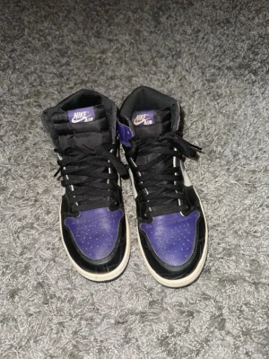 Nike Air Jordan 1 High Court Purple - Snygga Nike Air Jordan 1 High sneakers i färgerna lila, svart och vitt. Skorna har klassisk hög siluett, svart swoosh och vita detaljer. Ovandelen är i läder och de har svarta skosnören. Perfekta för dig som gillar streetwear och vill sticka ut.