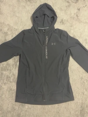Svart vindjacka från Under Armour - Snygg svart vindjacka från Under Armour med huva och hel dragkedja. Jackan har en jätte fin passform. Skön att ha på sig året runt. Pris kan diskuteras.