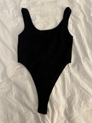 Svart ribbad body med djup rygg - Säljer en svart body med ribbad struktur och högt skuren benlinje. Bodyn har breda axelband och en djup rygg, vilket ger en snygg och modern look. Materialet är stretchigt och följsamt, perfekt för att matcha med jeans eller kjol.