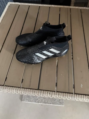 Svarta Adidas fotbollsskor utan snörning - Säljer ett par svarta Adidas fotbollsskor med silvriga ränder på sidan och utan snörning för en tight passform. Skorna har en strumpliknande design och är tillverkade i syntetmaterial som ger bra grepp och kontroll på planen. Perfekta för dig som vill ha stilrena och funktionella skor. Storlek 37