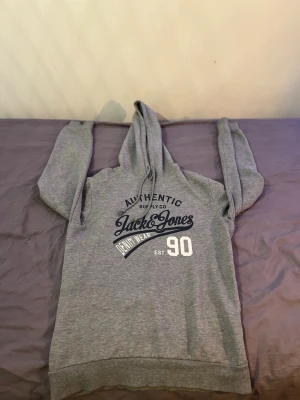 Grå hoodie från Jack & Jones - Snygg grå hoodie från Jack & Jones med tryck i marinblått och vitt på bröstet. Klassisk huva med dragsko och mjukt material, perfekt för en avslappnad stil. Stor logga och texten 'Denim Wear 90' framtill ger en sportig vibe.
