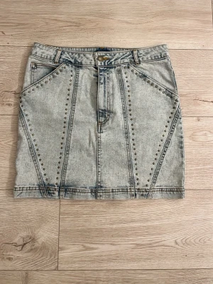 Kort jeanskjol med nitar - Snygg kort jeanskjol i ljusblå denim med coola nitar längs sömmarna framtill. Kjolen har fickor både fram och bak samt hällor för bälte. Perfekt för dig som gillar en edgy och trendig stil.