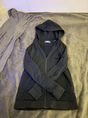 Svart zip hoodie från Jack and Jones. - En svart hoodie med dragkedja från Jack and Jones. Klassisk modell med huva och två stora fickor framtill. Tillverkad i mjukt bomullsmaterial som känns skönt mot huden. Perfekt för en avslappnad och stilren look.