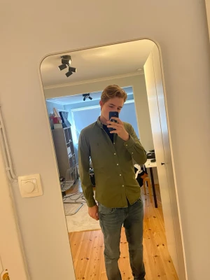 ÄKTA Olivgrön skjorta från Polo Ralph Lauren - Säljer en stilren olivgrön skjorta från Polo Ralph Lauren med klassisk button-down krage och vita knappar. Skjortan har lång ärm och diskret broderad logga på bröstet. Perfekt till jeans eller chinos för en clean look. Denna är 100% autentisk som man ser när jag skannat qr koden. Nästan aldrig använd pga den är lite liten på mig. Bara att höra av sig vid frågor!