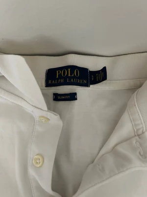 Vit slim fit piké från Polo Ralph Lauren - Vit pikétröja från Polo Ralph Lauren med klassisk krage, knappar framtill och broderad logga på bröstet. Slim fit-modell i mjukt bomullsmaterial. Perfekt för en clean och stilren look.