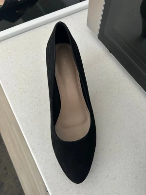 Svarta pumps i mocka-look - Stilrena svarta pumps med spetsig tå och låg klack. Skorna har en klassisk siluett och är klädda i ett material som liknar mocka. Insidan är ljusbeige och sulan är lätt vadderad för extra komfort. Perfekta för dig som gillar en tidlös och elegant stil.