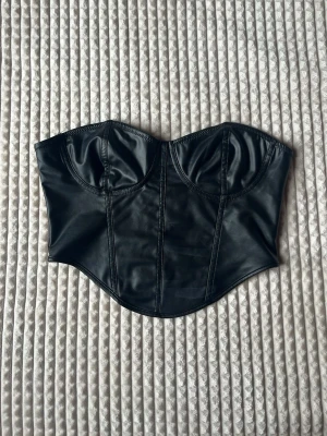 Svart bustierkorsett i läderlook - Snygg svart korsett med bustierform och markerade kupor. Materialet har en läderlook som ger en cool och edgy känsla. Korsetten har sömmar som framhäver formen och en böjd nederkant för en smickrande siluett. Perfekt för att skapa en statement-look.