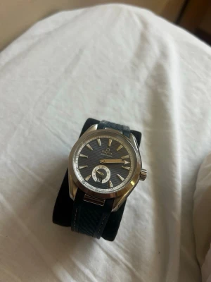 Omega klocka med blått läderband - Snygg Omega klocka med blått läderband och silverfärgat boett. Urtavlan är mörkgrå med randigt mönster och har tydliga indexmarkeringar samt en extra liten urtavla. Klassisk och stilren design som passar till många tillfällen.