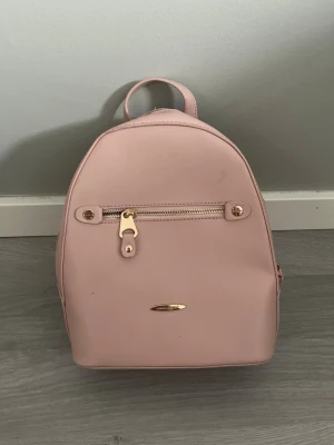 Ljusrosa ryggsäck med gulddetaljer - Söt ljusrosa ryggsäck i slät konstläder med guldfärgade dragkedjor och detaljer. Väskan har ett litet ytterfack framtill, justerbara axelband och ett praktiskt handtag upptill. Perfekt storlek för det viktigaste och ger en trendig touch till din outfit.
