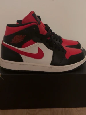 Air Jordan 1 Mid röd/svart/vit - Säljer ett par Air Jordan 1 Mid sneakers från Nike i färgerna röd, svart och vit. Skorna har klassisk Jordan-logga på sidan och tungan, svart snörning och en vit sula. Tillverkade i läder med snygga kontrastpaneler och retrovibe.