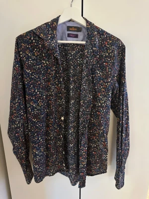 Blommig skjorta från Morris x Liberty - Mönstrad skjorta med småblommigt tryck i blått, vitt, rött och gult. Skjortan har klassisk krage, knäppning framtill och långa ärmar. Tillverkad i mjukt bomullstyg och har en avslappnad passform. Perfekt för dig som gillar färg och mönster.