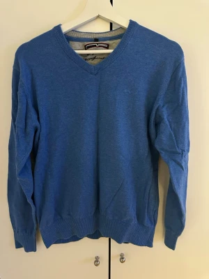 Blå stickad tröja med V-ringning - En stilren blå stickad tröja med V-ringad hals och ribbade muddar vid ärmslut och nederkant. Tröjan har en diskret broderad logga på bröstet och är tillverkad i ett mjukt material som passar perfekt till lager-på-lager. Långärmad och enkel att matcha.