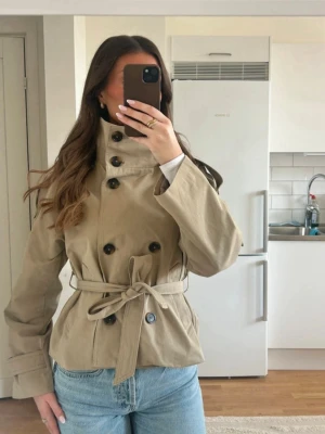 Kort beige trenchcoat med bälte bershka - Stilren beige trenchcoat med dubbelknäppning och högt krage. Jackan har ett matchande midjebälte och stora knappar framtill. Klassisk modell i kortare längd, perfekt till jeans. Materialet ser ut att vara bomull eller bomullsblandning. Jag kan tänka mig att byta mot en brun trenchcoat från bershka i storlek xs eller s. Lånade bilder!