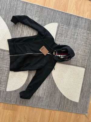 Svart hoodie från Burberry - Snygg svart hoodie från Burberry med dragkedja och klassiskt rutigt foder i huvan. Hoodien har fickor framtill och justerbar dragsko i huvan. Tillverkad i mjukt material som känns skönt mot huden. Perfekt för dig som vill ha en stilren och exklusiv look.