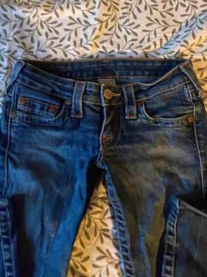True religion jeans - Snygga mörkblå jeans med slitningar och dekorativa knappar på fickorna. Klassisk femficksmodell med orangea sömmar och låg midja. Perfekta till en avslappnad streetstyle-look. Väldigt lowwaist och de är bootcut. Fina överallt förutom att det finns en defekt (se sista bilden)!! 💓💓
