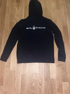 Svart hoodie från Sail Racing - Svart hoodie med stor huva och Sail Racing-logga tryckt i vitt på bröstet. Klassisk rak passform med långa ärmar och ribbade muddar. Perfekt för en avslappnad och sportig stil.