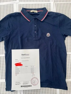 Marinblå pikéskjorta från Moncler - Snygg marinblå pikéskjorta från Moncler med klassisk krage och knappar framtill. Kragen har röda och vita ränder som ger en sportig touch. Moncler-logga på bröstet. Perfekt till jeans eller shorts för en clean look.