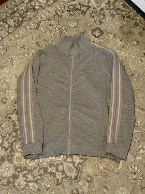 Grå ziptröja från iets frans... - Säljer en grå ziptröja från iets frans... med hög krage och hel dragkedja framtill. Tröjan har ribbade muddar och två ljusare ränder längs båda ärmarna för en sportig look. Perfekt för en avslappnad stil.