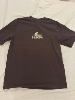 Brun Natural Reserve t-shirt - Säljer en brun t-shirt från Natural Reserve med tryck fram och stort motiv på ryggen med fåglar och text. T-shirten har normal passform och är kortärmad. Perfekt för dig som gillar naturinspirerade prints och avslappnad stil. Det står Loose S, så mer en M eller oversize S