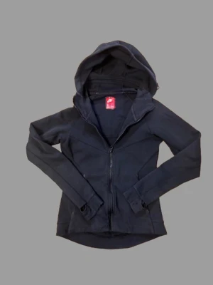 Svart Nike tech från Nike med dragkedja - Snygg svart hoodie från Nike i storlek XS. Hoodien har dragkedja framtill, långa ärmar och en stor huva. Tillverkad i mjukt och stretchigt material som känns bekvämt. Perfekt för dig som gillar sportig och clean stil.