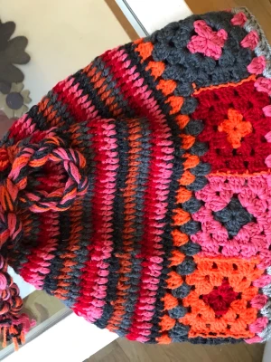 Färgglad virkad mössa med tofs - Unik virkad mössa i starka färger som rött, orange, rosa och grått. Mössan har ränder och mormorsrutor samt en stor tofs upptill. Tillverkad i mjukt garn, perfekt för dig som vill sticka ut med en retro och lekfull stil.