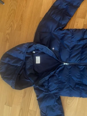 Mörkblå dunjacka med huva från GANT - Snygg mörkblå dunjacka från GANT med huva och dragkedja framtill. Jackan har en klassisk quiltad design och diskret logga på bröstet. Perfekt för kalla vinterdagar och riktigt skön att bära tack vare det lätta dunmaterialet.