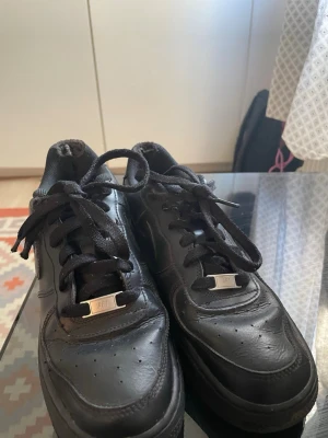 Svarta Nike Air Force 1 sneakers - Klassiska helsvarta Nike Air Force 1 sneakers i läder med lågt skaft och svarta skosnören. Swoosh-logga på sidan och metallbricka med 'AF1' på snörningen. Perfekta för en clean och stilren look gamla.