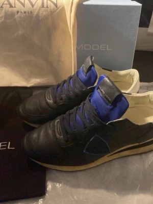 Philippe Model svarta sneakers med blå detaljer - Snygga svarta sneakers från Philippe Model med blå detaljer på plös och sidor. Skorna har en klassisk siluett, svarta skosnören och vit insida. Yttermaterialet är skinn och sulan är beige. Perfekta för dig som gillar stilrena och trendiga sneakers. Hör av dig vid funderingar.