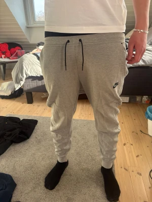 Nike tech - Grå mjukisbyxor från Nike i storlek L. Stilren design med avsmalnande ben, resår i bensluten och justerbar dragsko i midjan för perfekt passform. Klassisk logga på benet som ger en sportig och trendig känsla.  Sköna och mjuka i materialet – perfekta för både vardag, träning eller hemmamys. Lätta att matcha med hoodie, t-shirt eller jacka. I fint skick.