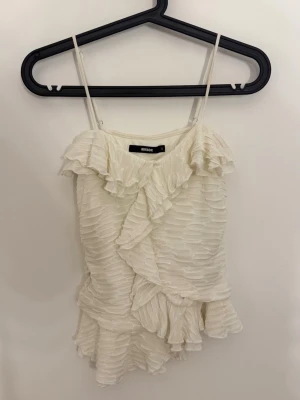 Vit volangtop från Bikbok - Supersöt vit topp från Bikbok med tunna axelband och volangdetaljer. Toppen har ett plisserat tyg och flera lager volanger som ger en romantisk vibe. Perfekt att styla med jeans eller kjol för en trendig look.
