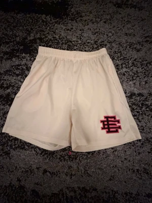 Basket shorts - Säljer ett par beiga shorts med elastisk midja och en broderad röd och svart logga på vänster ben. Shortsen har en avslappnad passform och är perfekta för basket och sommaren . Materialet känns mjukt och lätt, ett sportigt material.
