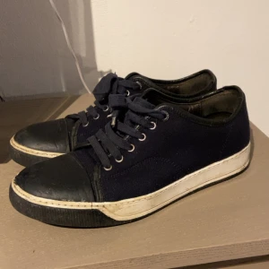 Lanvin  - Säljer ett par mörkblå låga sneakers med tå och sula i svart gummi och ovandel i tyg. Skorna har svarta skosnören och metallöljetter. Klassisk och enkel design som passar till det mesta.