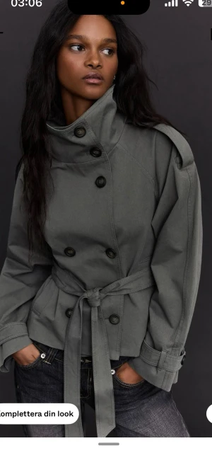 Grå trenchcoat med knytskärp - Snygg grå trenchcoat med dubbelknäppning, höga axelpartier och knytskärp i midjan. Jackan har bred krage, långa ärmar med justerbara muddar och två sidofickor. Perfekt för att ge din outfit en cool touch.