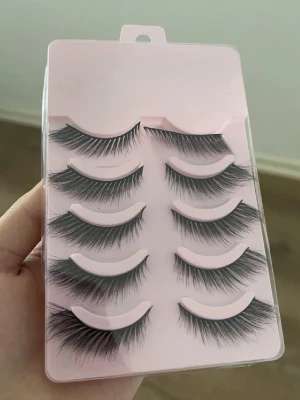 Svarta volymfransar i 5-pack - Säljer ett set med fem par svarta lösögonfransar med volym och fluffig design. Fransarna har en naturlig böj och sitter på en tunn, flexibel remsa. Förpackningen är ljusrosa och genomskinlig. Perfekt för att skapa en intensiv ögonlook.