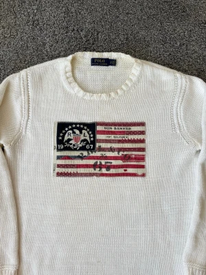 Vit stickad tröja Polo Ralph Lauren - Vit stickad tröja från Polo Ralph Lauren med amerikansk flagga och textdetaljer på bröstet. Tröjan har rund halsringning, långa ärmar och ribbade muddar. Materialet ser ut att vara bomull och passformen är klassisk.