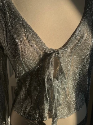 Silvrig meshkofta med knytband - Trendig silvrig meshkofta med lång ärm och djup v-ringning. Koftan har ett tunt, glittrigt material och knyts framtill med band. Perfekt för att lägga till lite extra bling till din outfit.